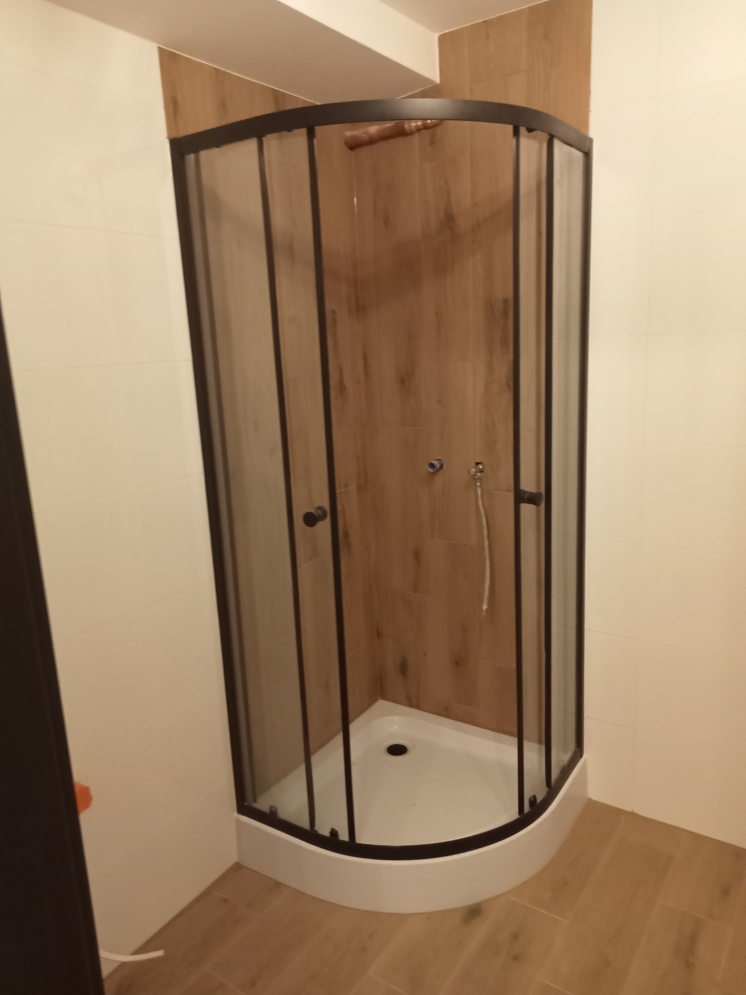 Kabina prysznicowa z czarną ramą i drewnopodobnymi płytkami ściennymi, widoczne przyłącza wodne i biały brodzik