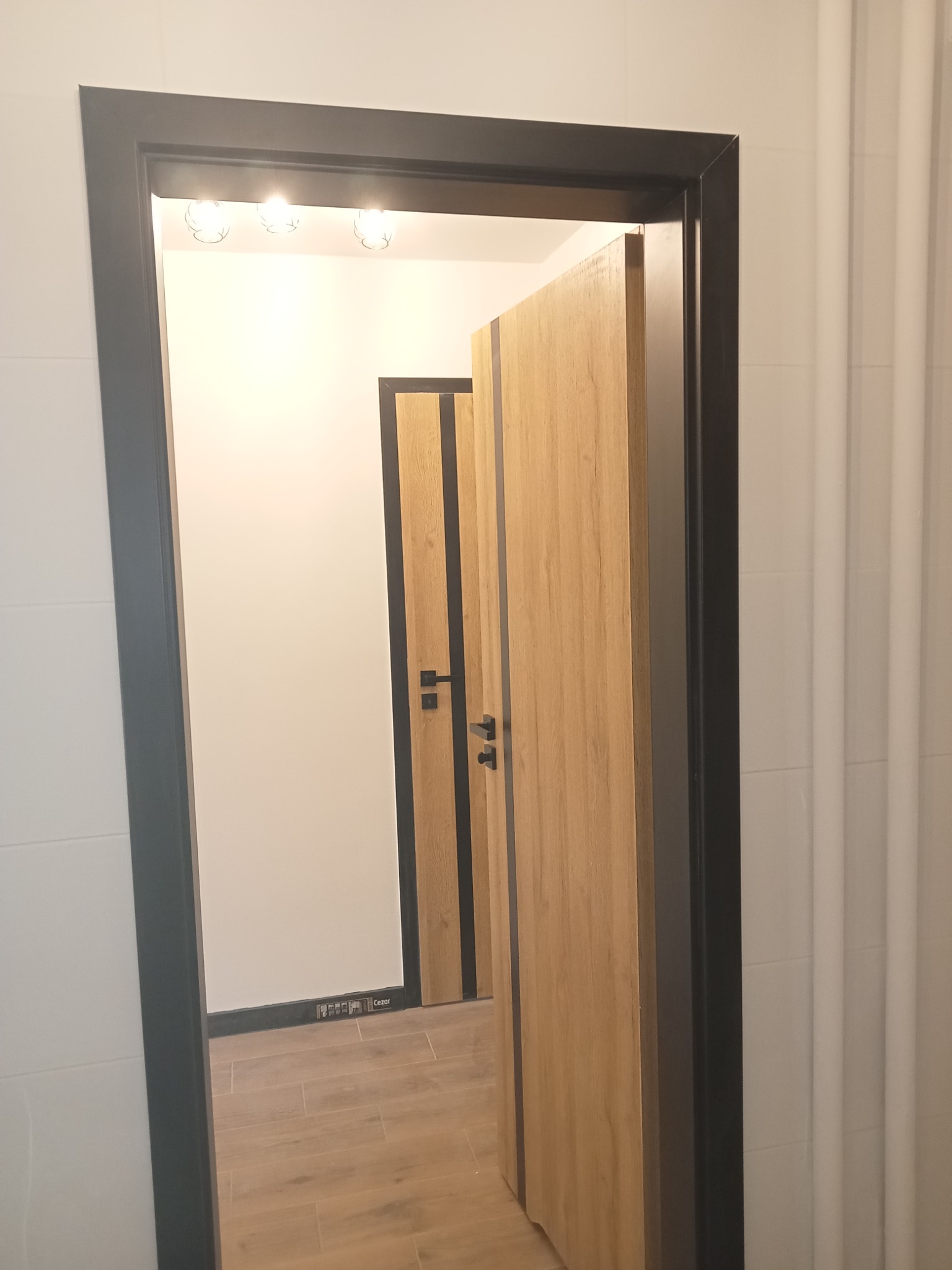 Widok na wnętrze przez otwarte drzwi z czarną ościeżnicą, ukazujący podłogę z paneli, białe ściany i kolejne drzwi w głębi, również z czarnymi akcentami.