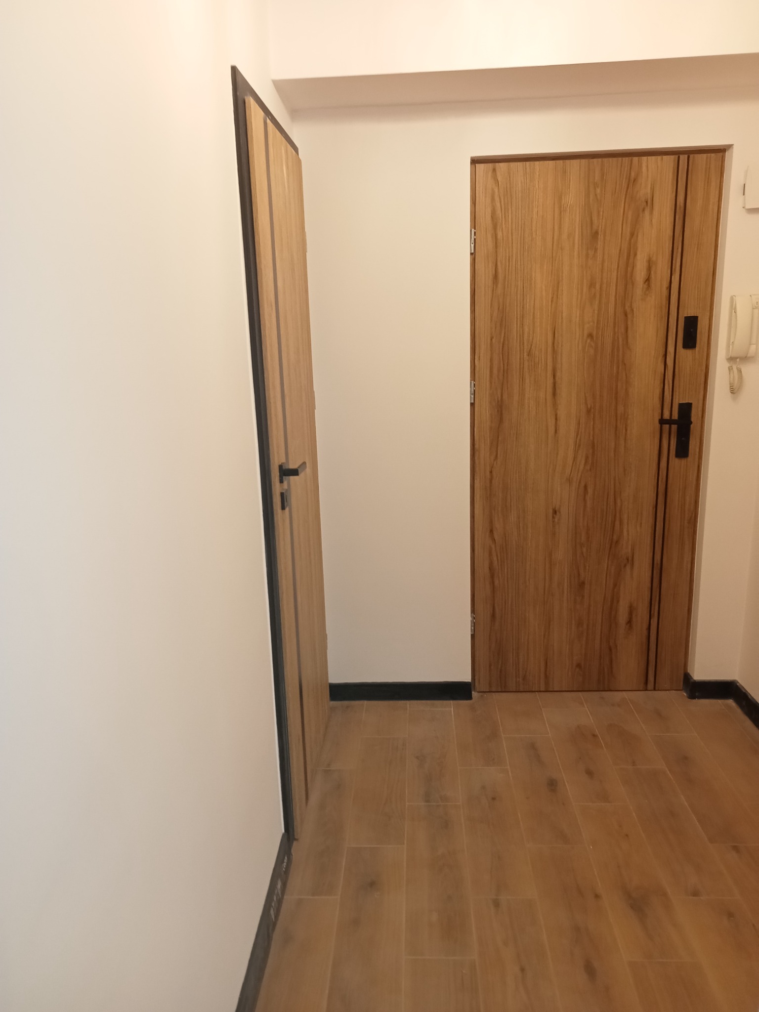 Dwa drewnopodobne drzwi wewnętrzne z czarnymi detalami, widoczne na tle białych ścian i drewnopodobnej podłogi z płytek.