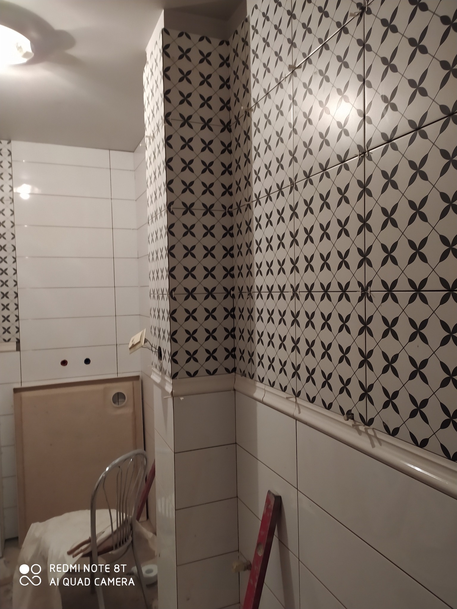 Świeżo ułożone, wzorzyste płytki w łazience, z widocznymi krzyżykami dystansowymi i poziomnicą, w tle kabina prysznicowa i krzesło.