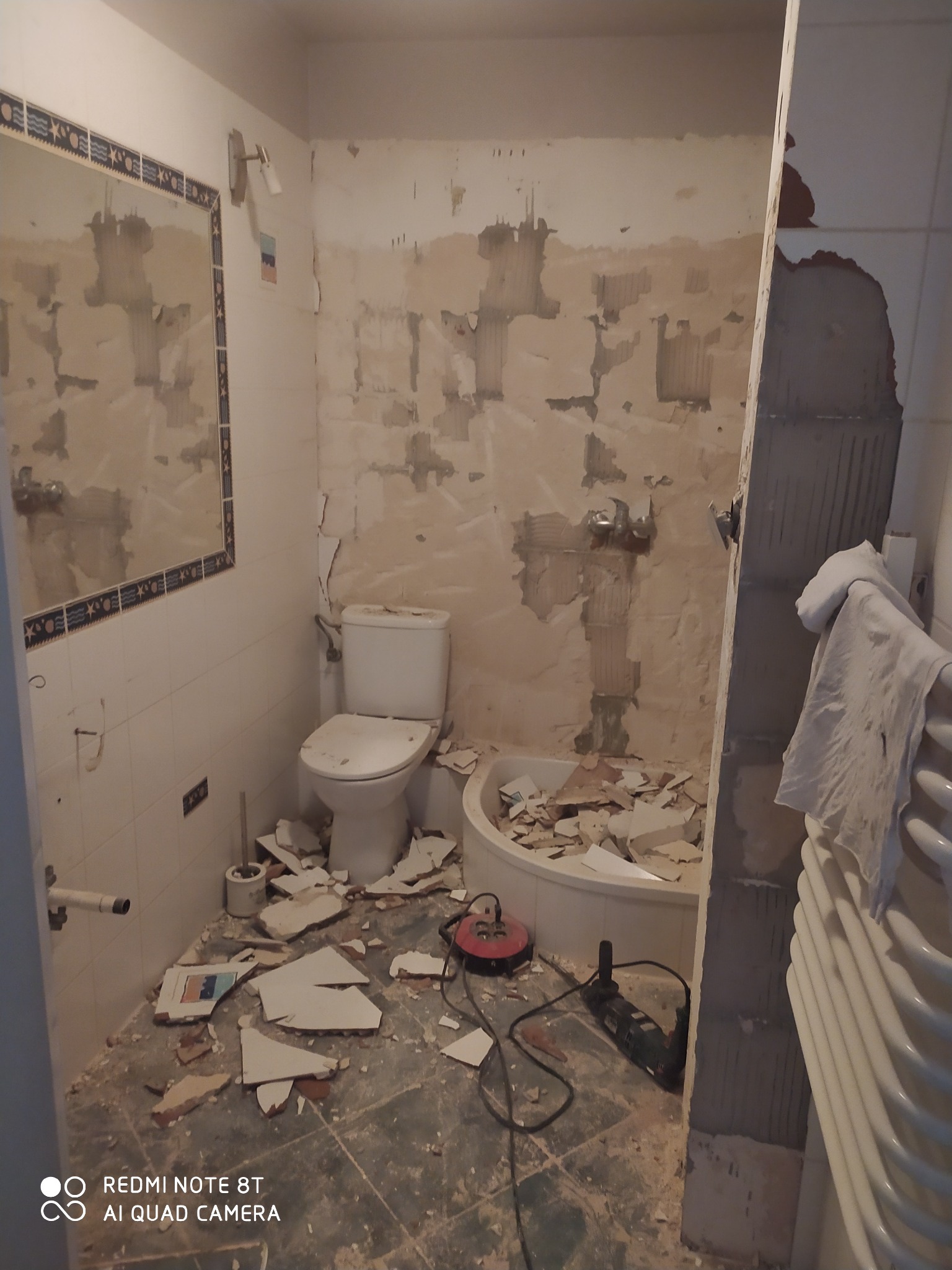 Łazienka w trakcie remontu: zerwane płytki, odsłonięte ściany, gruz na podłodze, widoczny sedes, wanna i narzędzia budowlane.