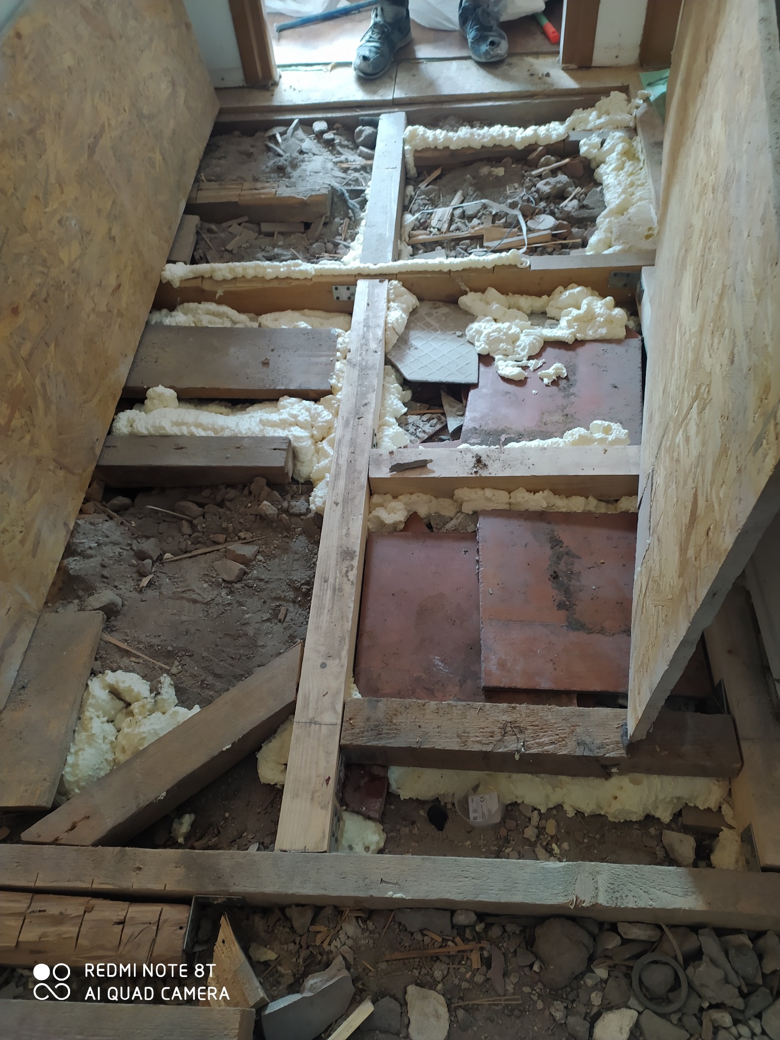 Podłoga w trakcie remontu: widoczne drewniane legary, izolacja pianką montażową, fragmenty starej posadzki i płyty OSB, buty robocze w tle.