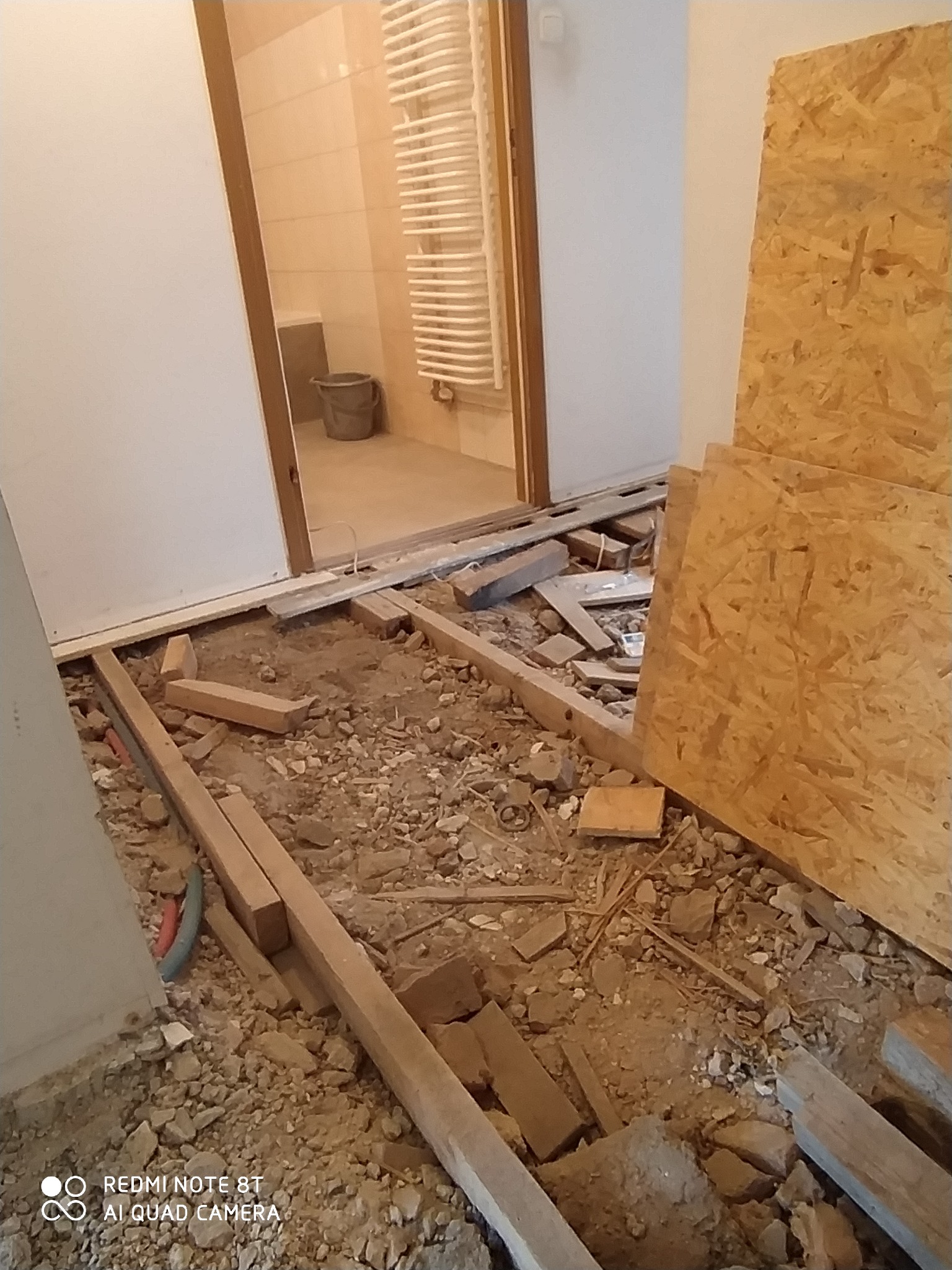 Przygotowanie podłoża pod nową podłogę w łazience, widoczne legary, gruz i płyty OSB, w tle widoczna łazienka z grzejnikiem drabinkowym.