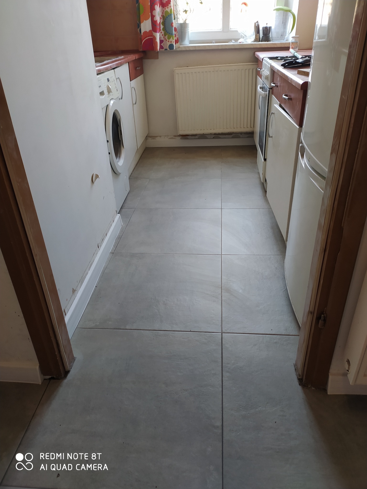 Ułożone szare płytki podłogowe w wąskiej kuchni w Myślenicach, widok z perspektywy wejścia, z widoczną pralką i fragmentem lodówki po bokach.