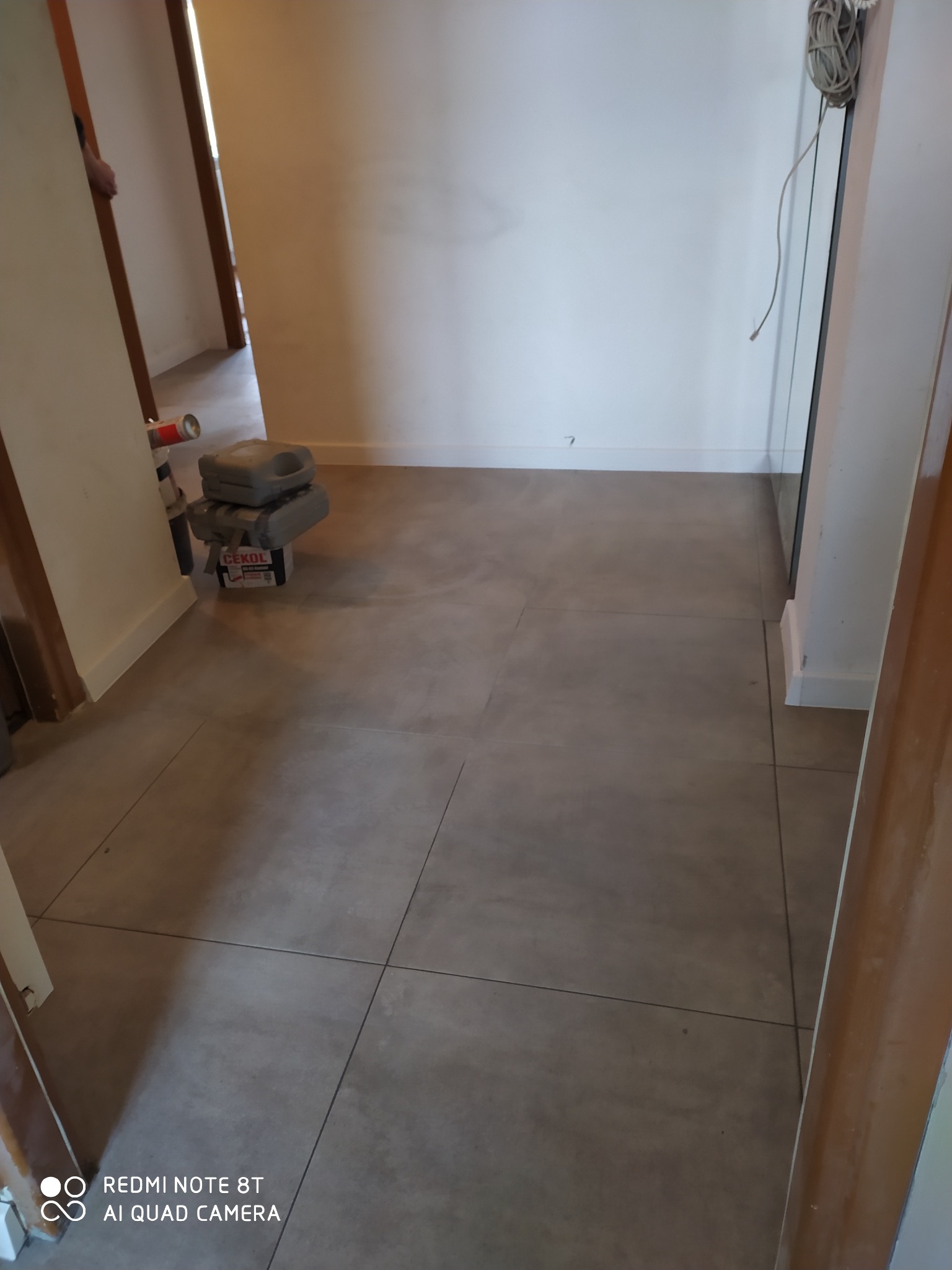 Korytarz z szarymi, kwadratowymi płytkami podłogowymi; widoczny fragment ściany, drzwi i narzędzia pracy w tle.