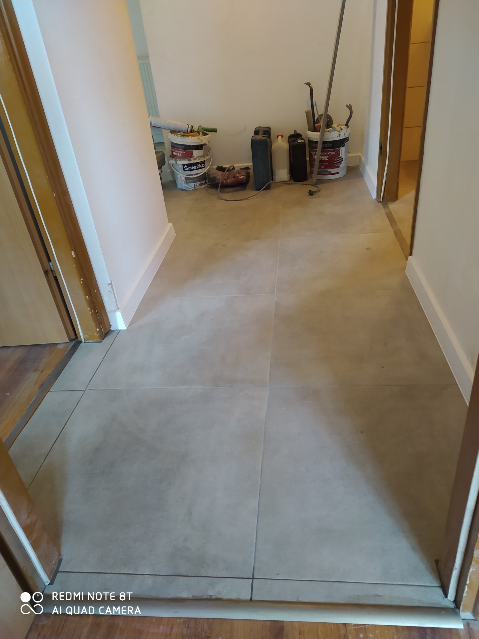 Korytarz z nowymi, beżowymi płytkami podłogowymi; w tle widoczne wiadra z farbą, puszki i narzędzia malarskie, sugerujące świeżo zakończone prace.