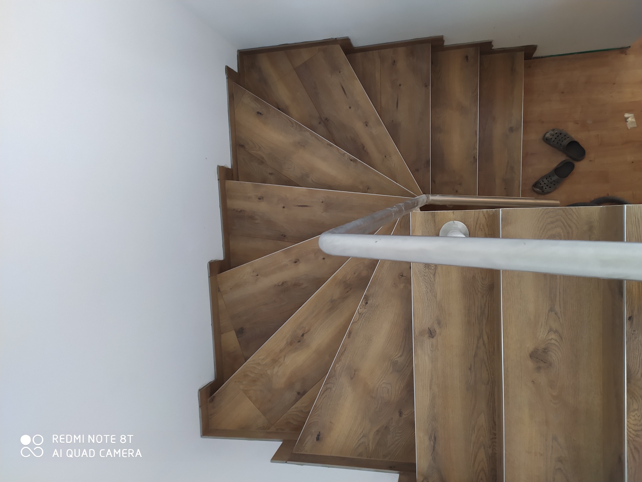 Widok z góry na drewniane, spiralne schody z metalową poręczą, obok leżą kapcie, widoczny fragment podłogi w trakcie remontu.