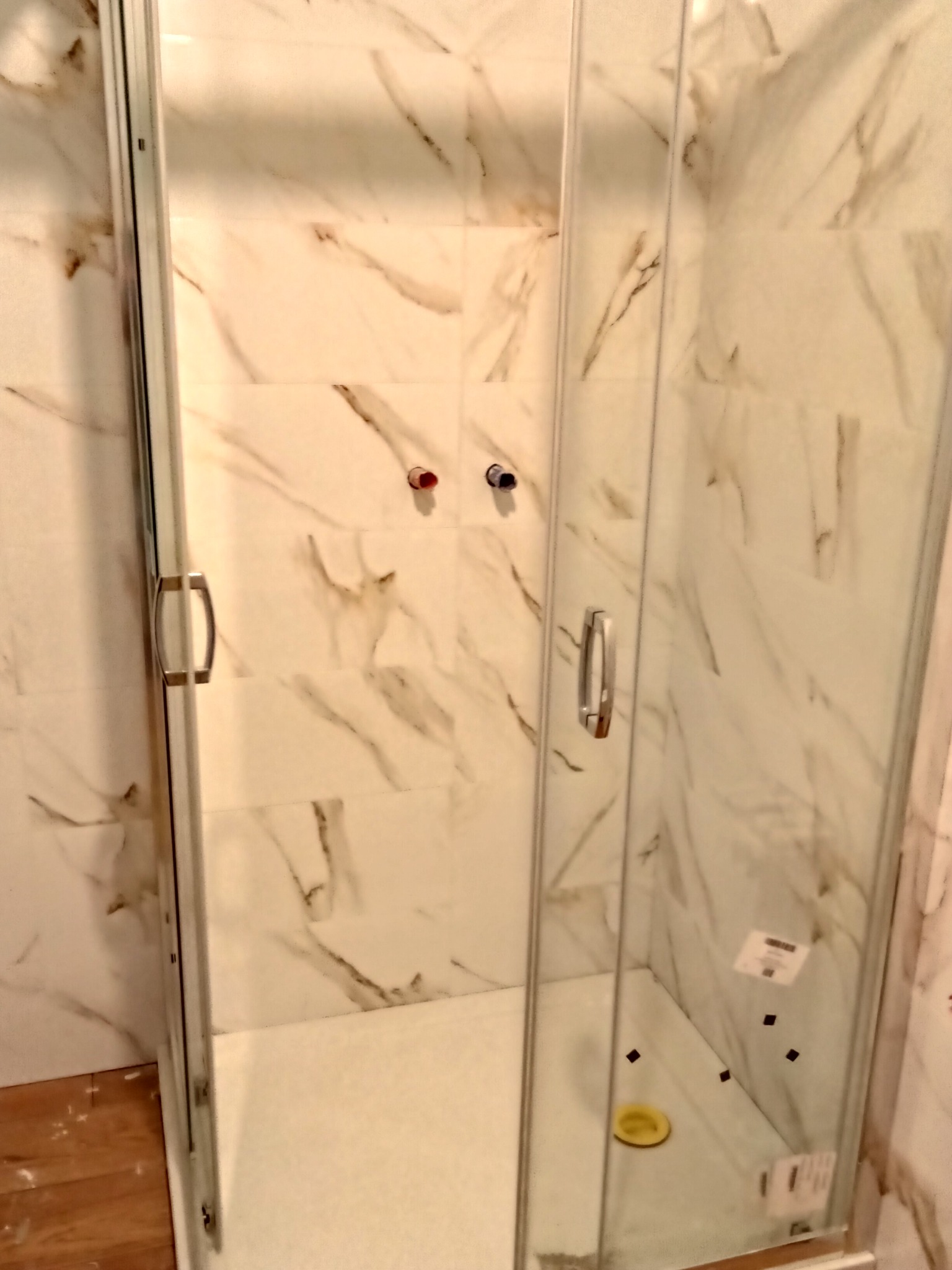 Szklana kabina prysznicowa z przesuwnymi drzwiami, wykończona płytkami imitującymi marmur, widoczne metalowe uchwyty i biały brodzik.