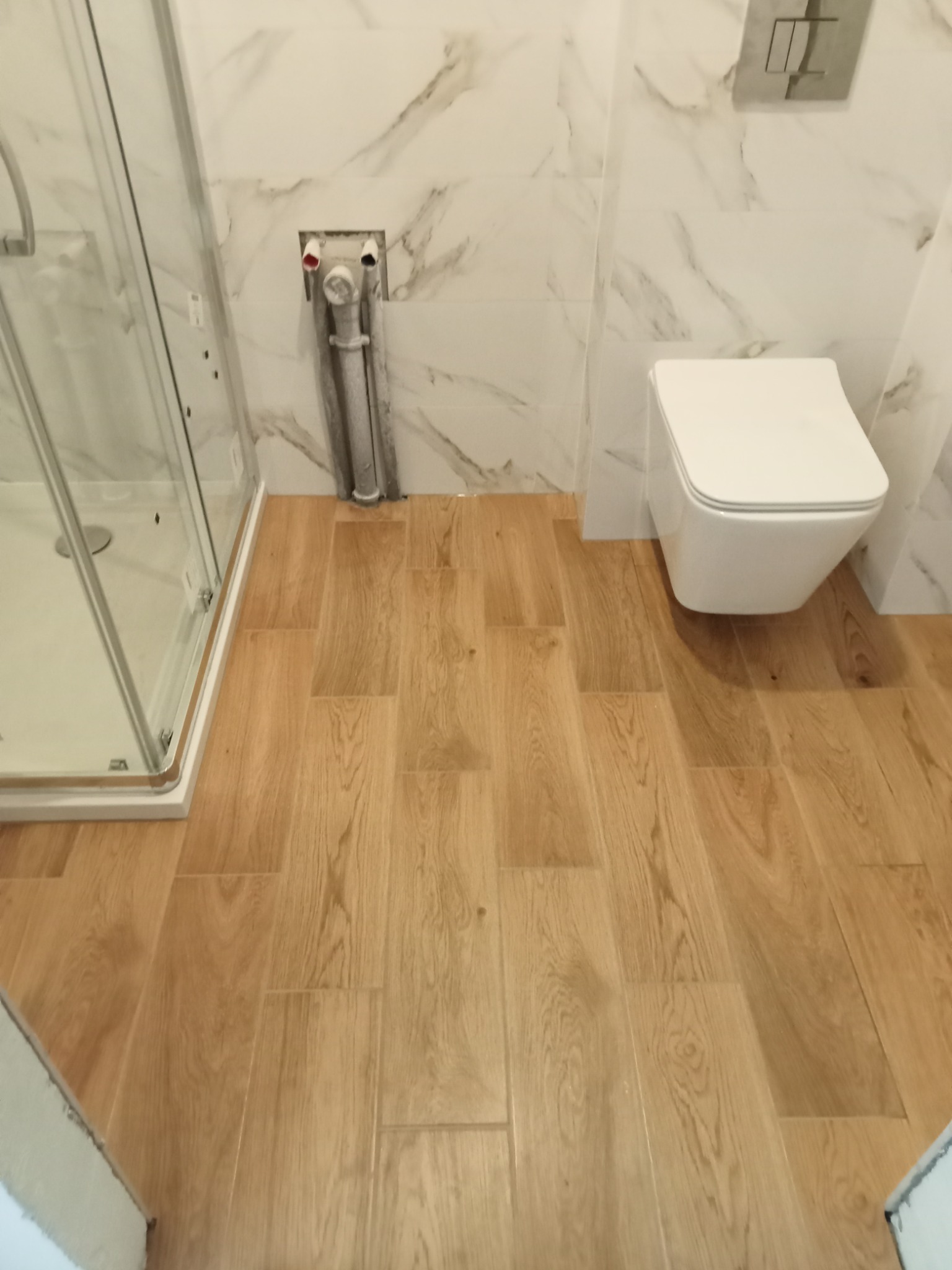 Nowoczesna łazienka z drewnopodobną podłogą, marmurowymi płytkami ściennymi, wiszącą toaletą i kabiną prysznicową z chromowanymi detalami; widoczne niezamaskowane rury instalacyjne.