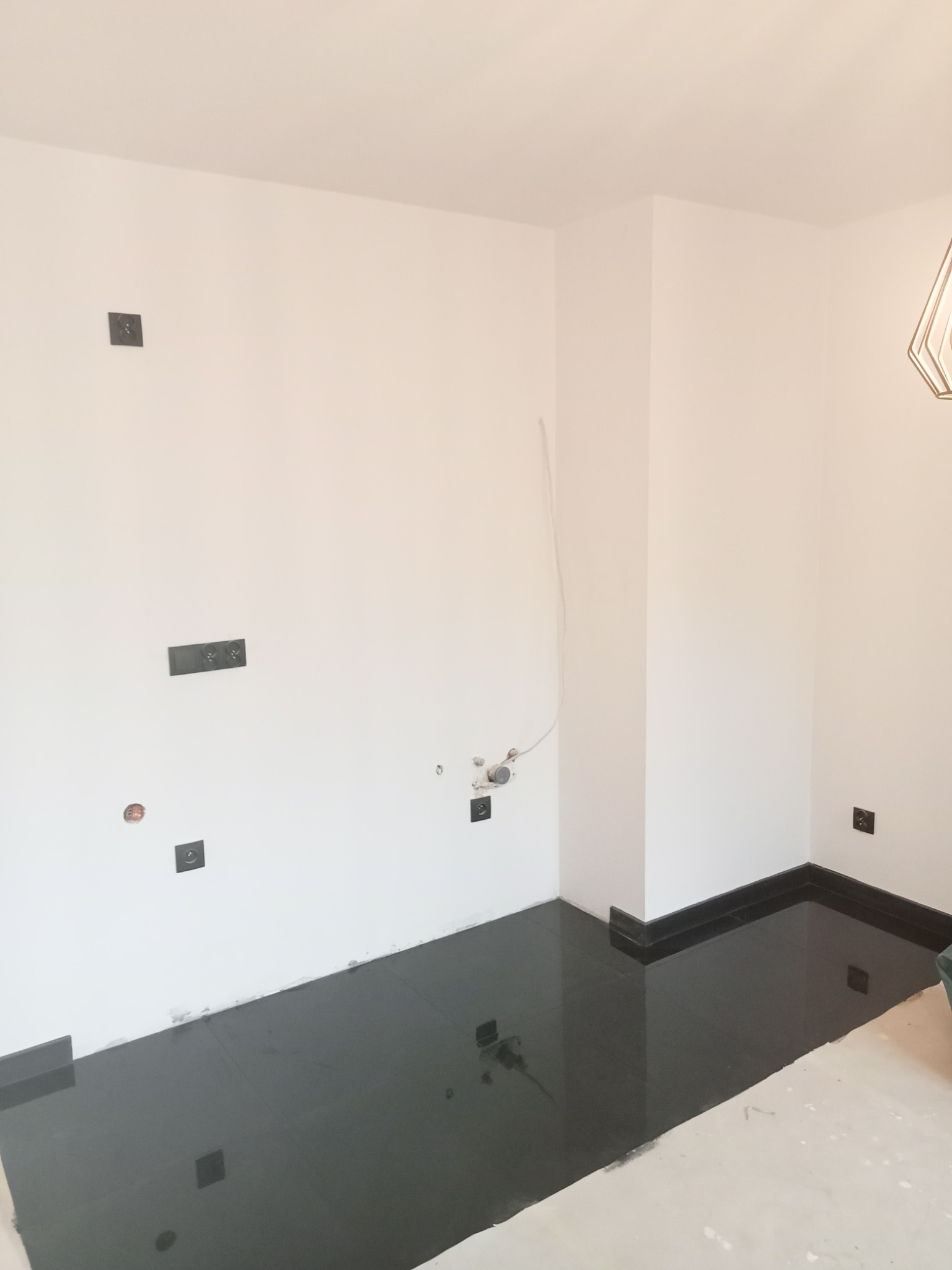 Świeżo ułożona, błyszcząca, czarna podłoga z płytek w rogu pomieszczenia z białymi ścianami, widoczne gniazdka i instalacje elektryczne, brak listew przypodłogowych.