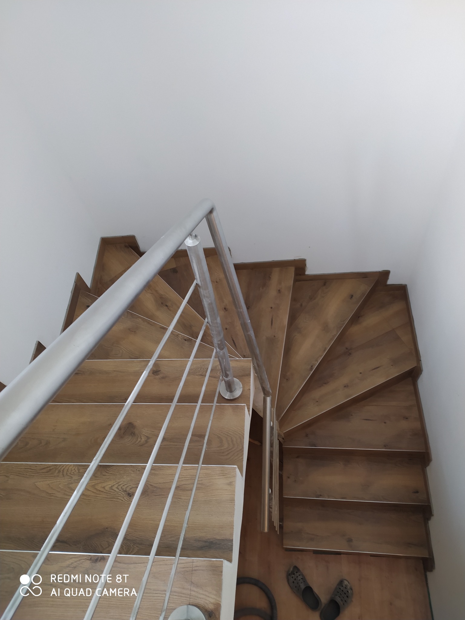 Widok z góry na drewniane schody spiralne z metalową balustradą, widoczne klapki i odkurzacz na dole schodów.