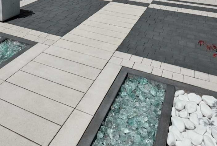 Szachownica z ciemnoszarej i jasnoszarej kostki brukowej, z wkomponowanymi rabatami wypełnionymi szkłem i białymi kamieniami.