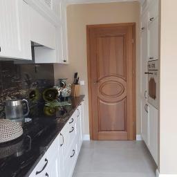 Tilmon Damian Piaseczny - Wykończona kuchnia w stylu klasycznym: białe szafki z czarnymi uchwytami, czarny blat z granitu, drewniane drzwi z owalnym panelem, szara podłoga z płytek.