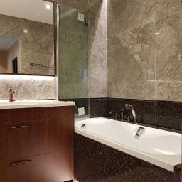 Tilmon Damian Piaseczny - Nowoczesna łazienka z marmurowymi płytkami, drewnianą szafką pod umywalką i wanną obudowaną mozaiką.