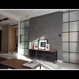 Tilmon Damian Piaseczny - Minimalistyczny salon z szarą ścianą, czarnymi, przeszklonymi przesuwnymi drzwiami i metalową szafką pod ścianą, oświetlony popołudniowym słońcem.