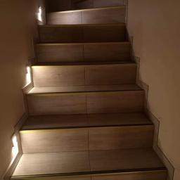 Tilmon Damian Piaseczny - Wykończone schody wewnętrzne wyłożone płytkami imitującymi drewno z subtelnym oświetleniem LED w ścianie.