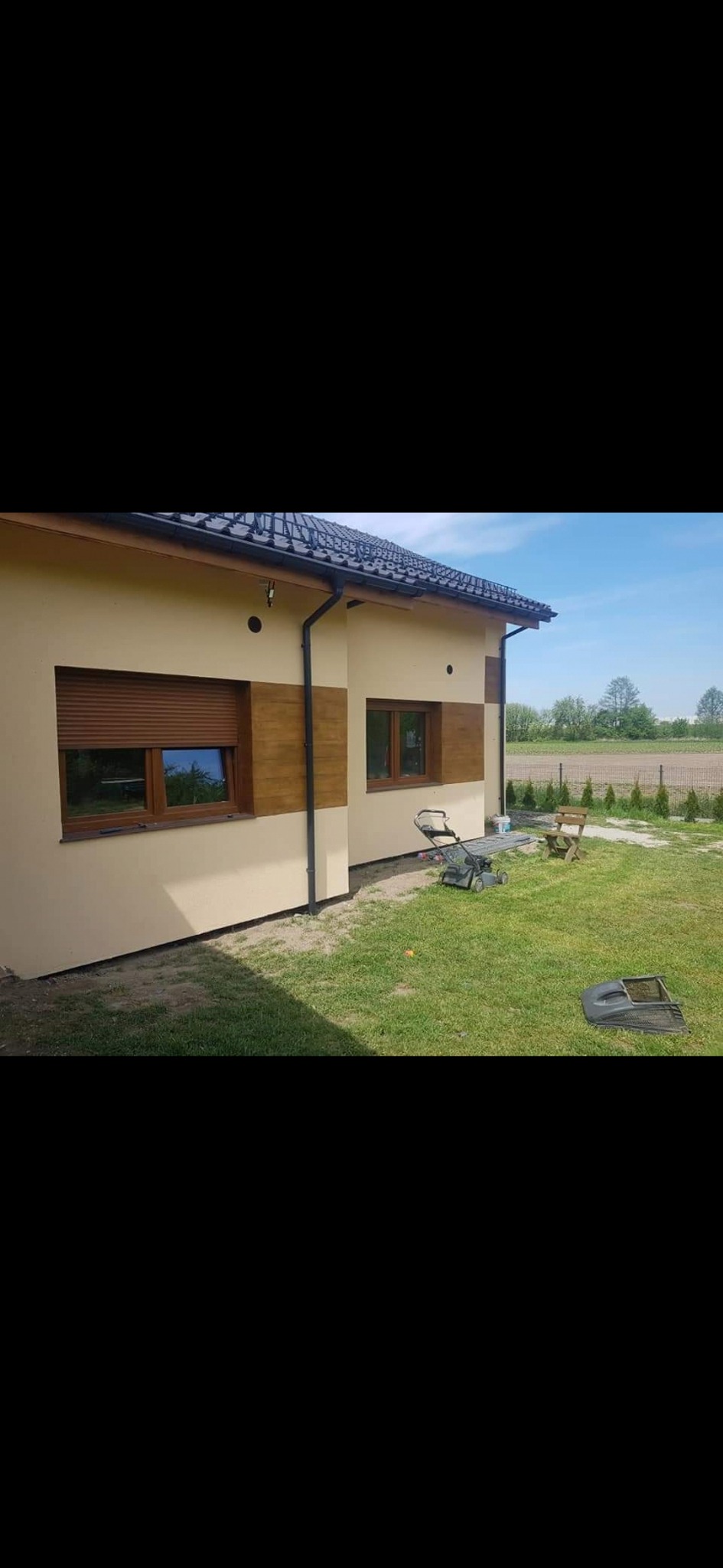 Elewacja domu jednorodzinnego w jasnym odcieniu z drewnopodobnymi panelami dekoracyjnymi wokół okien, widoczna kosiarka na trawniku.