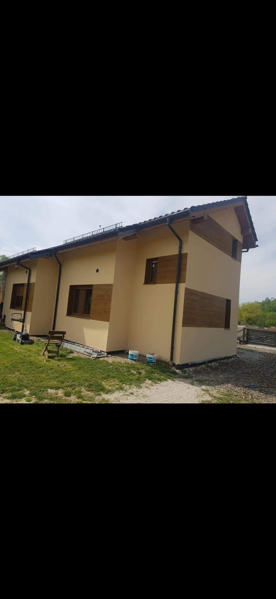 Dwu-kondygnacyjny budynek szeregowy z beżową elewacją i poziomymi pasami imitującymi drewno, widoczne dwa wiadra z farbą, trawnik i częściowo wykończony teren wokół.