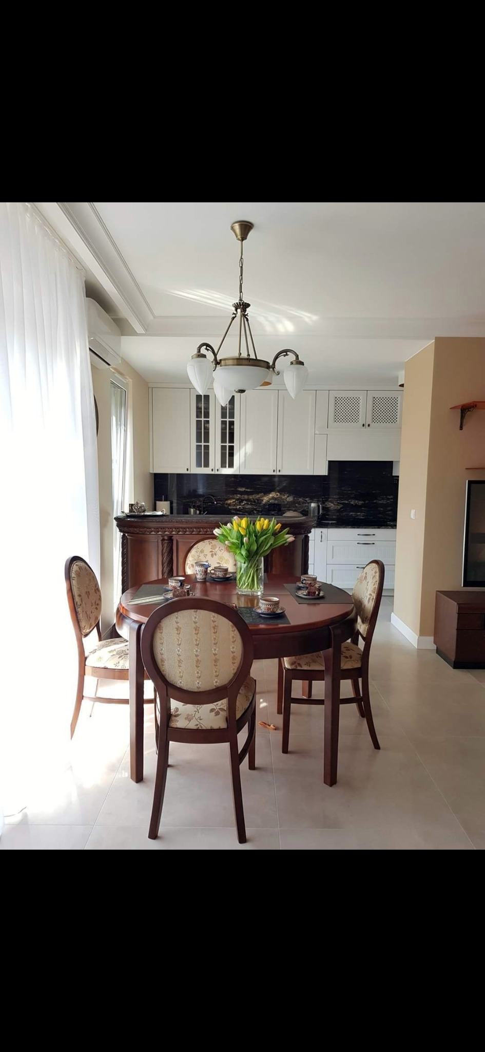 Jasne wnętrze jadalni z okrągłym, drewnianym stołem i krzesłami z tapicerką w kwiaty, na stole wazon z żółtymi tulipanami i filiżanki, w tle biała zabudowa kuchenna z czarnym blatem.