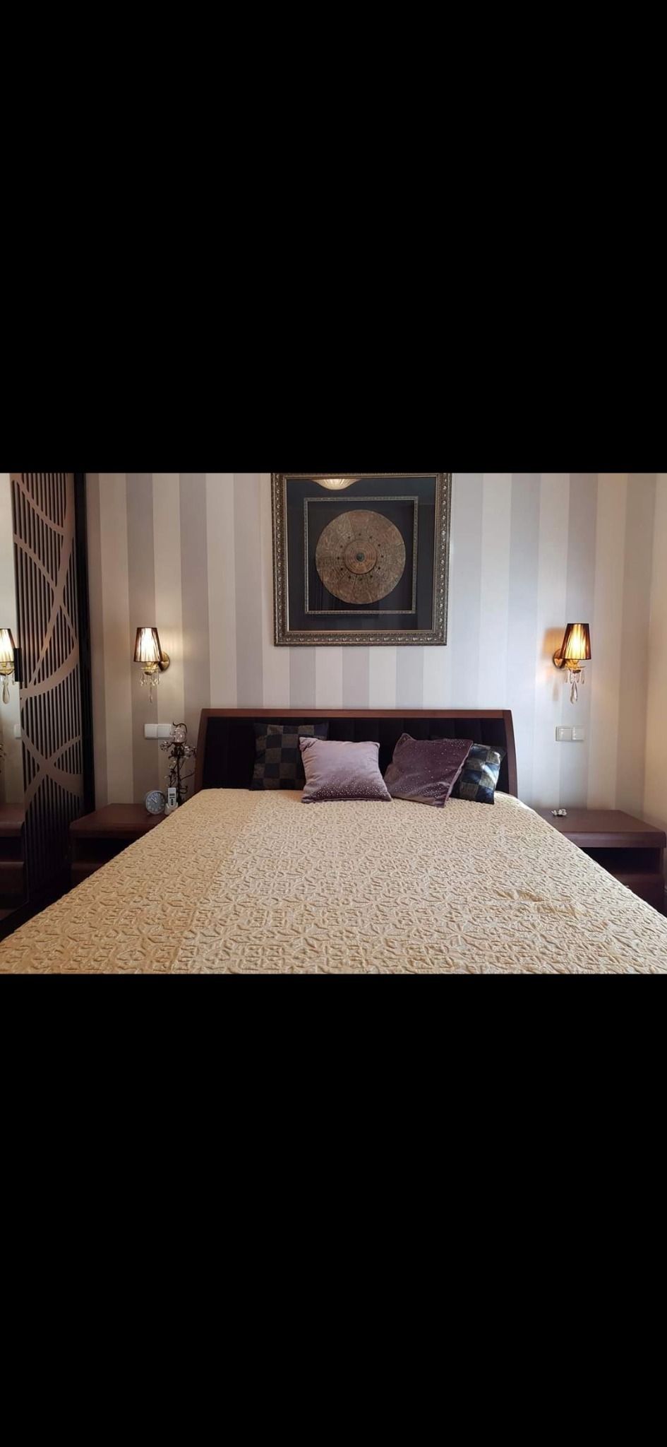 Sypialnia z łóżkiem małżeńskim, wzorzystą narzutą, fioletowymi poduszkami i lampkami nocnymi z abażurami, na ścianie obraz w ozdobnej ramie.