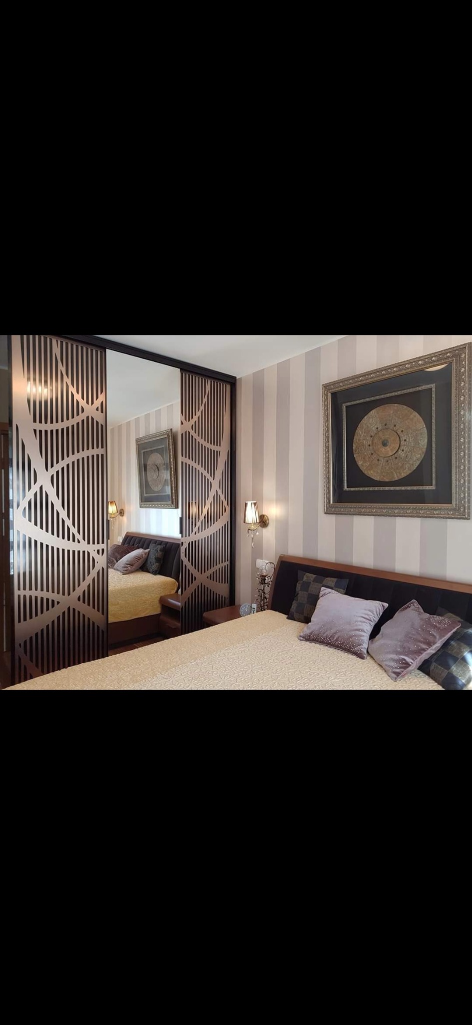 Sypialnia z łóżkiem małżeńskim, lampkami nocnymi i przesuwnymi drzwiami szafy z dekoracyjnym ażurem, odbijającymi się w lustrze.