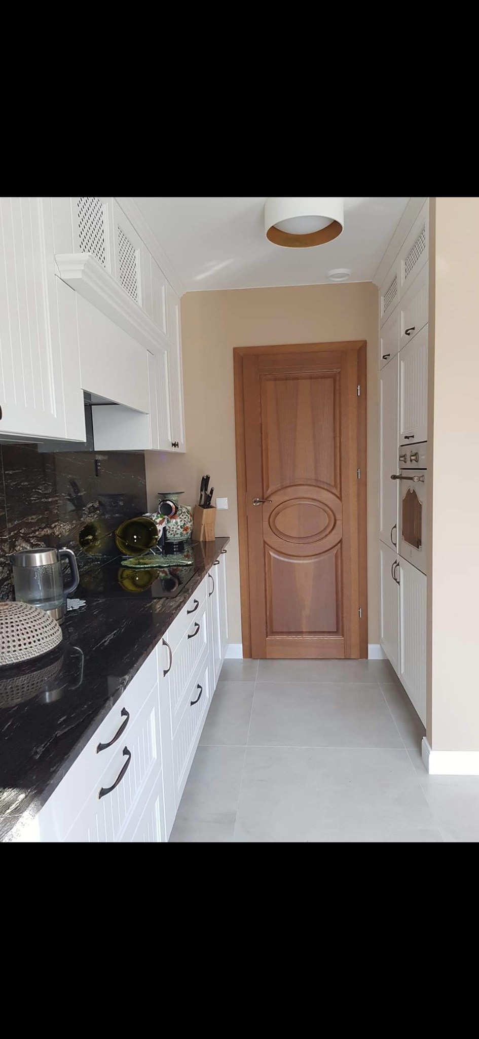 Wykończona kuchnia w stylu klasycznym: białe szafki z czarnymi uchwytami, czarny blat z granitu, drewniane drzwi z owalnym panelem, szara podłoga z płytek.