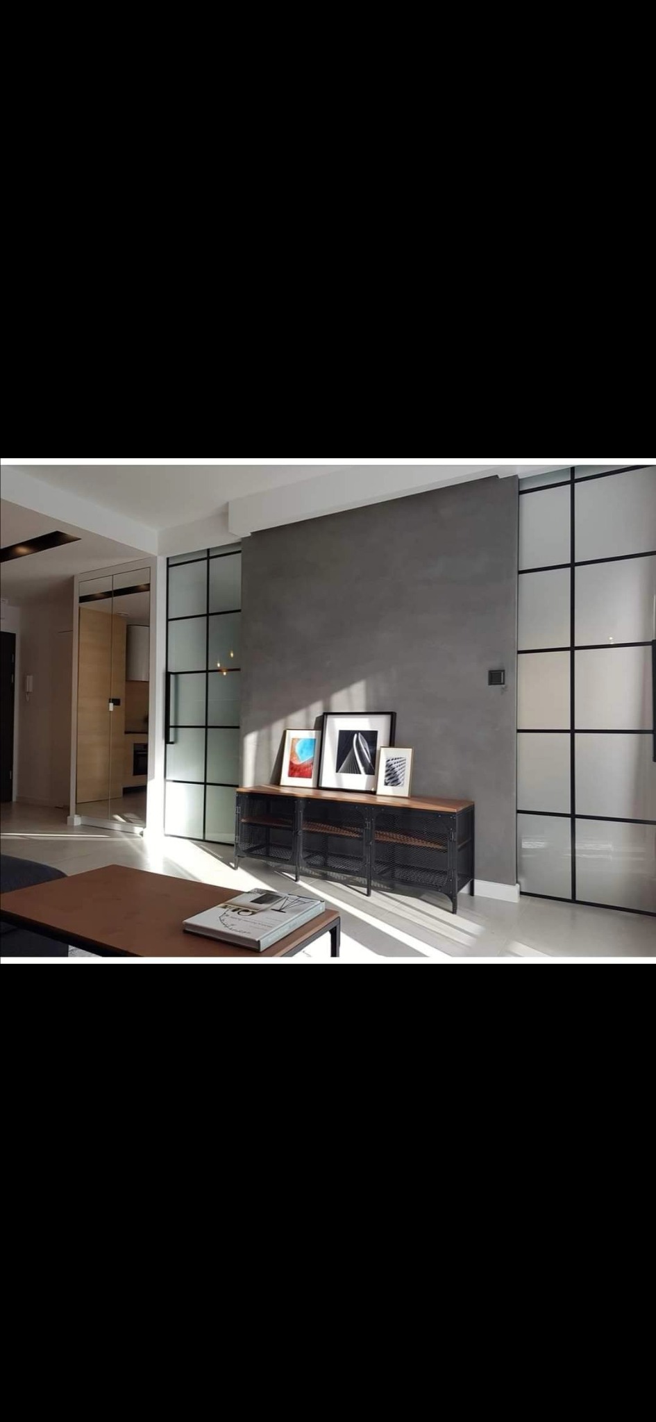 Minimalistyczny salon z szarą ścianą, czarnymi, przeszklonymi przesuwnymi drzwiami i metalową szafką pod ścianą, oświetlony popołudniowym słońcem.