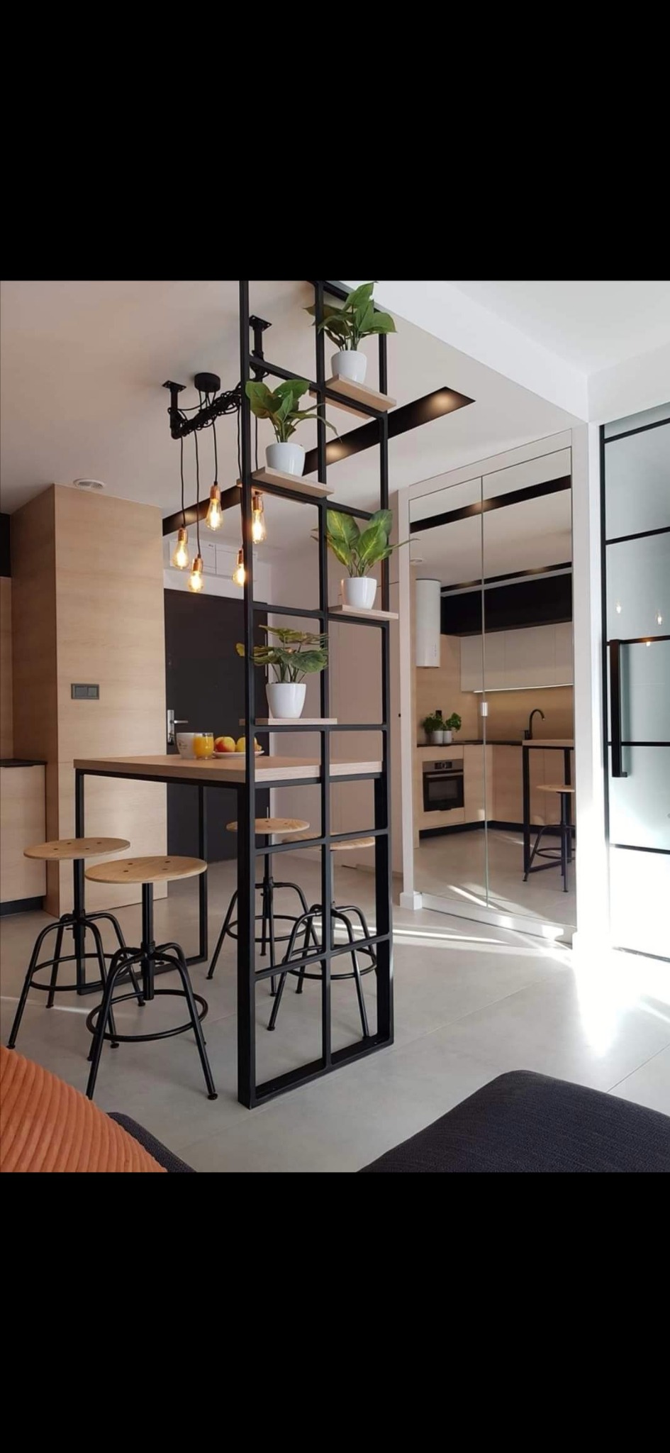 Nowoczesna aranżacja wnętrza z industrialnym regałem oddzielającym strefę jadalną od kuchni, akcenty roślinne na półkach, drewniane stołki barowe i wiszące lampy z odsłoniętymi żarówkami.