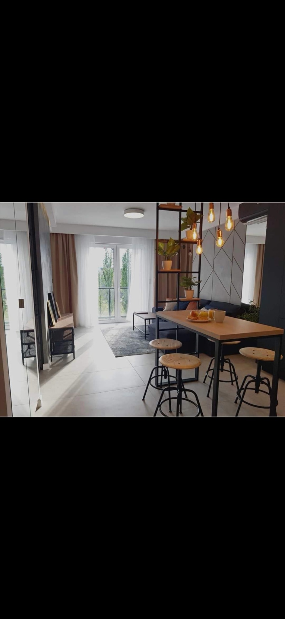 Jasne wnętrze salonu z drewnianym stołem barowym i czterema stołkami, widok na okno balkonowe z zielenią, oświetlenie punktowe i geometryczna ściana w tle.