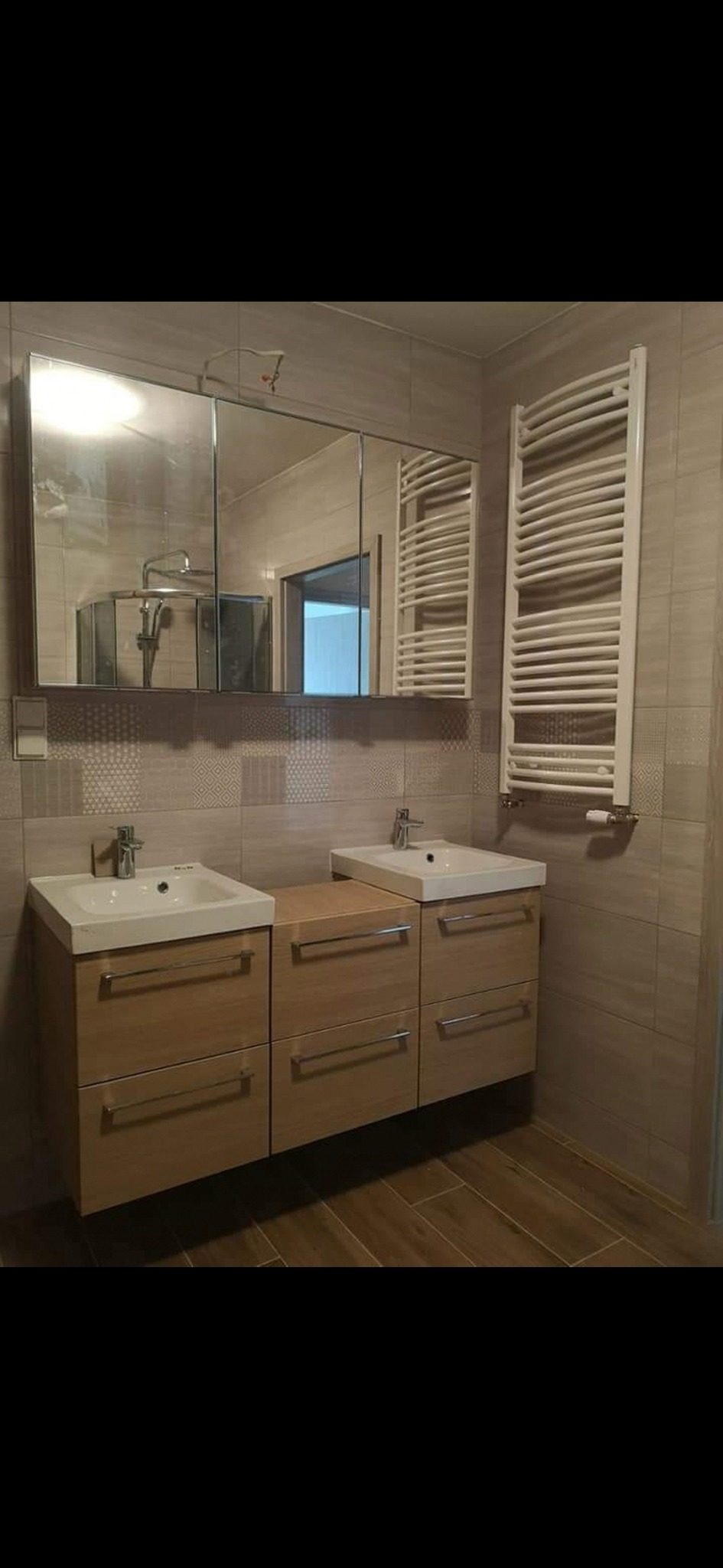 Nowoczesna łazienka z dwoma umywalkami na drewnianych szafkach, lustro z szafką i grzejnik drabinkowy na ścianie wyłożonej beżowymi płytkami z subtelnym wzorem.