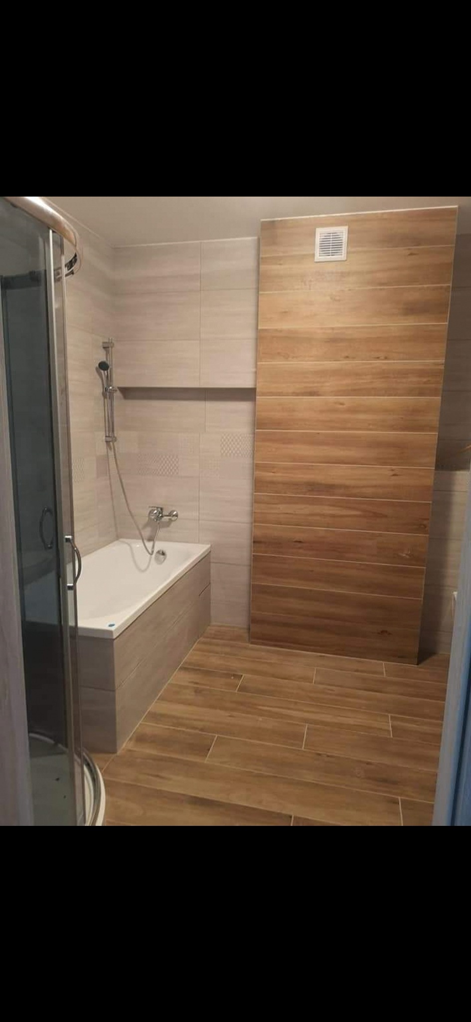 Wykończona łazienka z kabiną prysznicową, wanną obudowaną płytkami i ścianą wyłożoną płytkami imitującymi drewno, z kratką wentylacyjną.