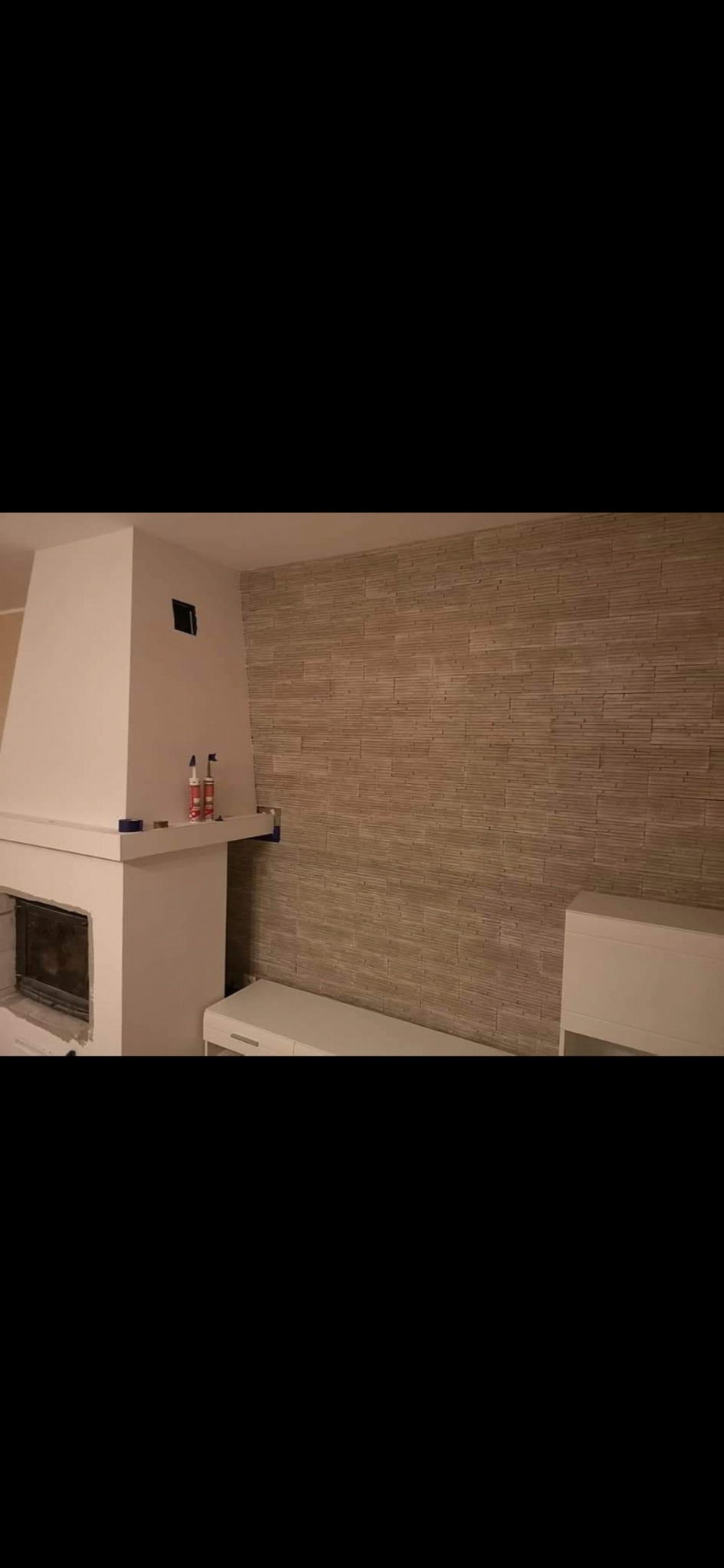 Ściana wykończona dekoracyjną okładziną imitującą kamień, kominek w trakcie wykończenia, widoczne tuby z klejem i poziomica.