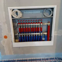 Rozdzielacz ogrzewania podłogowego w białej obudowie, widoczne rury niebieskie i czerwone, instalacja na folii grzewczej z siatką, ściana w stanie surowym.