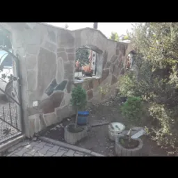 Kamienny mur z oknami, częściowo otynkowany, z elementami dekoracyjnymi z kutego żelaza, otoczony zielenią i roślinami w betonowych donicach. Widoczne ślady prac wykończeniowych.