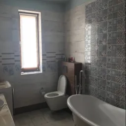 Jasna łazienka z wanną na ozdobnych nóżkach, toaletą wiszącą i mozaiką na ścianie, oświetlona światłem dziennym z okna.