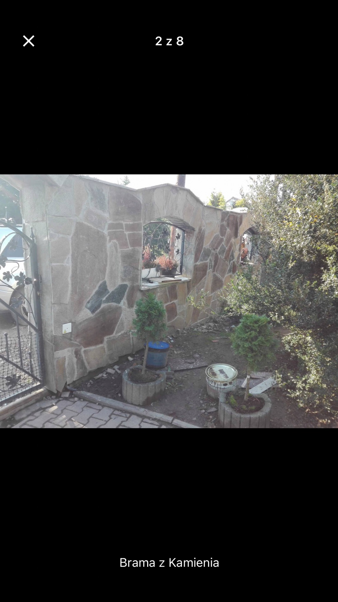 Kamienny mur z oknami, częściowo otynkowany, z elementami dekoracyjnymi z kutego żelaza, otoczony zielenią i roślinami w betonowych donicach. Widoczne ślady prac wykończeniowych.