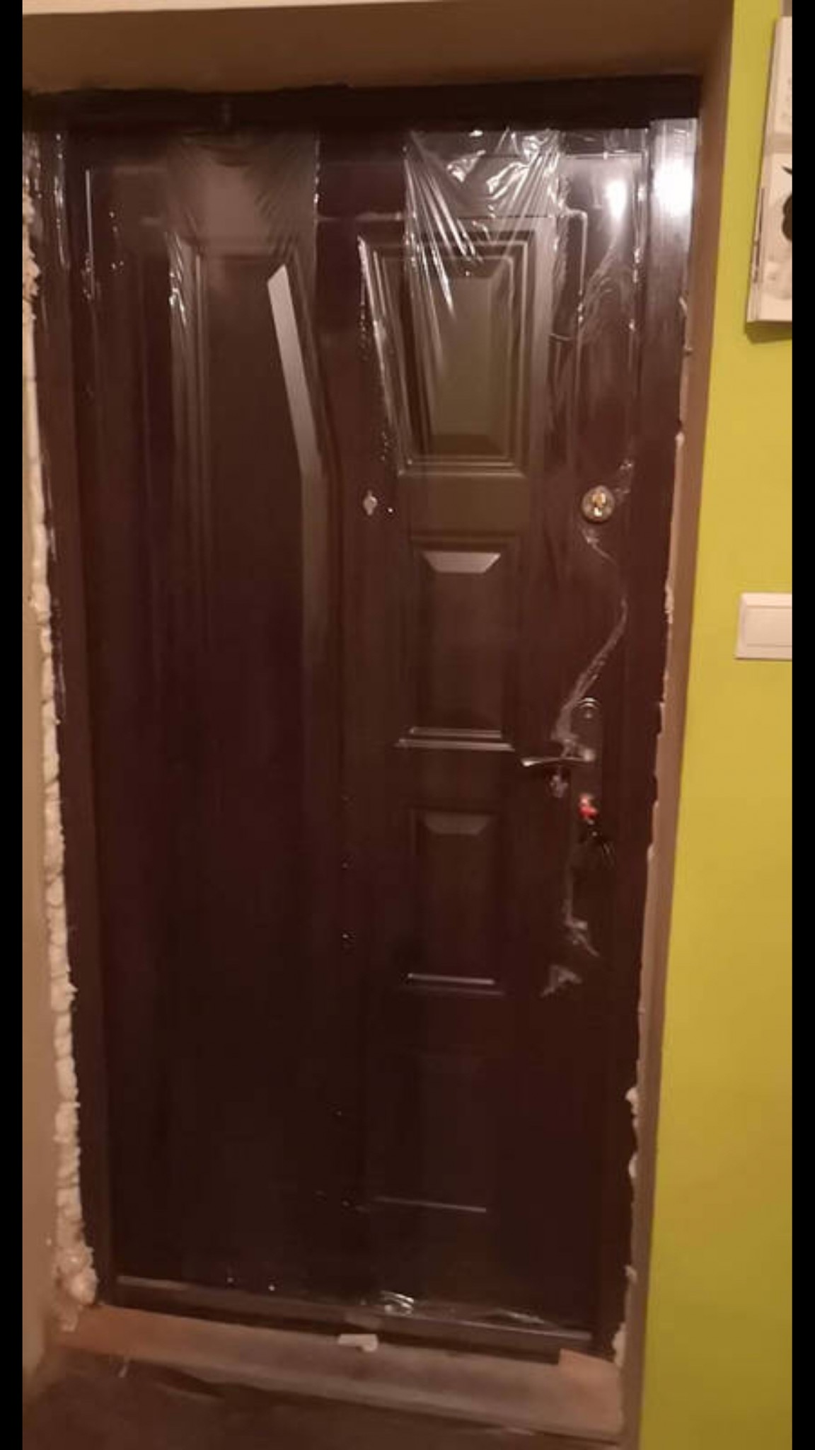 Nowa, brązowa, metalowa drzwi wejściowe, jeszcze w folii ochronnej, zamontowane w ościeżnicy z widoczną pianką montażową, na tle żółtej ściany.