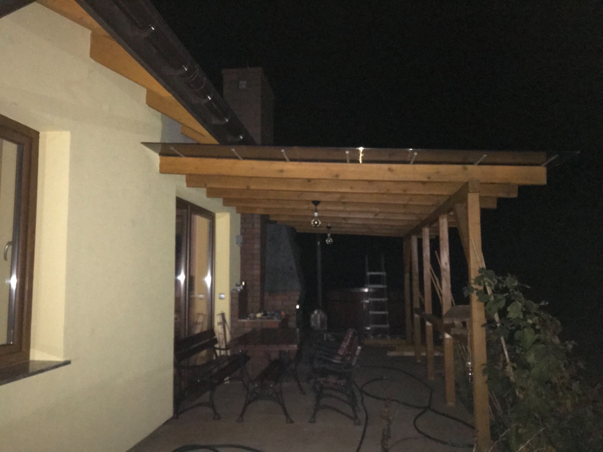 Zadaszona drewniana konstrukcja tarasu przylegająca do budynku z oknami i drzwiami, widoczna w nocy, z ciemnymi meblami ogrodowymi i roślinnością w tle.