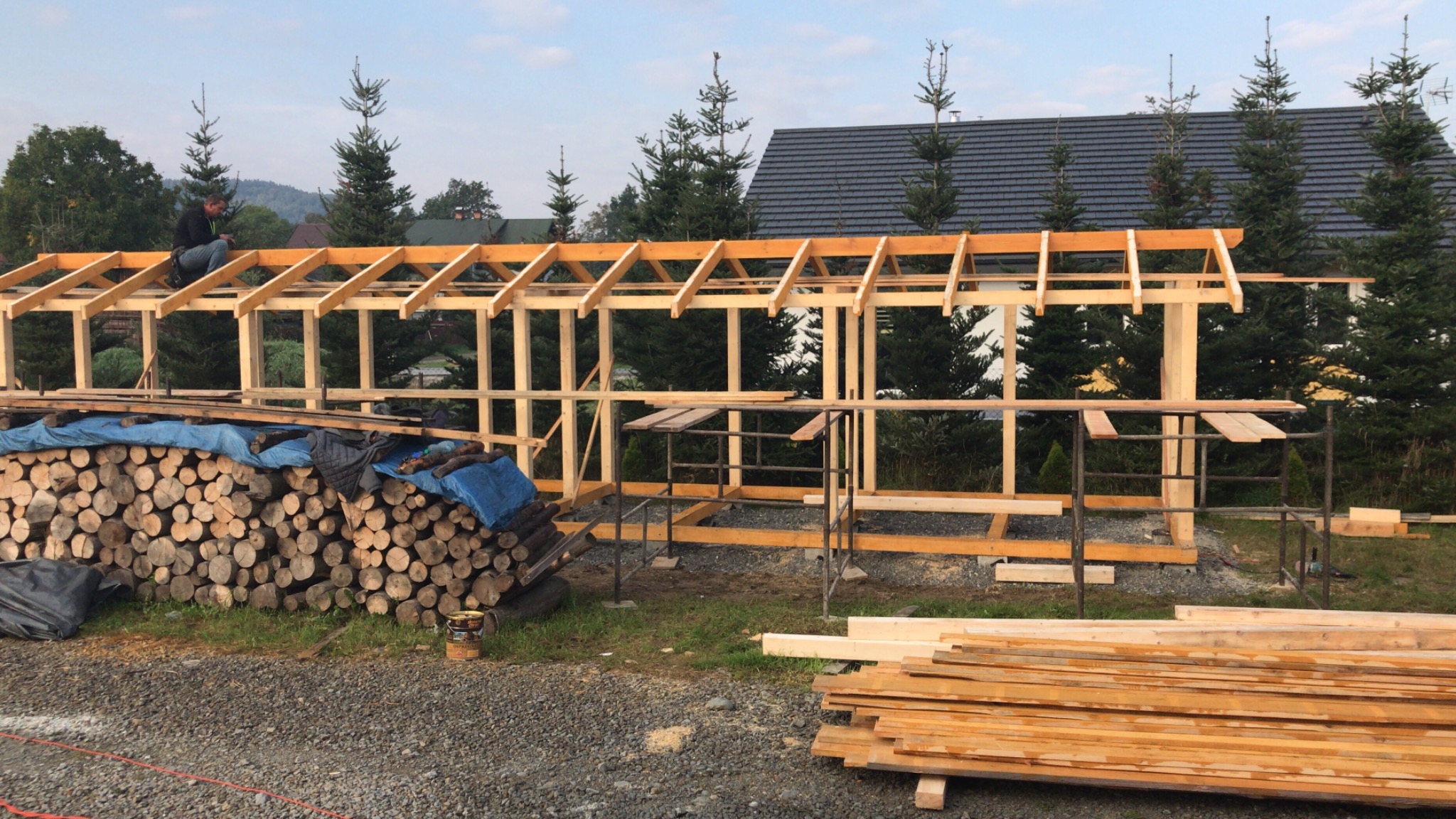 Konstrukcja drewniana z belek, prawdopodobnie zadaszenie balkonu lub tarasu, w trakcie budowy, z widocznym pracownikiem na górze i stosem drewna obok, na tle drzew iglastych i domu z ciemnym dachem.