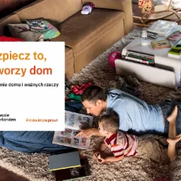 Ojciec z synem leżą na dywanie w salonie, oglądając książkę z obrazkami; w tle sofa, stolik kawowy z zabawkami i pufy; widoczny fragment reklamy towarzystwa ubezpieczeniowego z hasłem 'Ubezpiecz to...