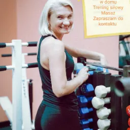 Uśmiechnięta kobieta w sportowym stroju stoi przy stojaku z hantlami w siłowni, na ścianie kartka z napisem 'Trening personalny w domu, Trening siłowy, Masaż, Zapraszam do kontaktu'.
