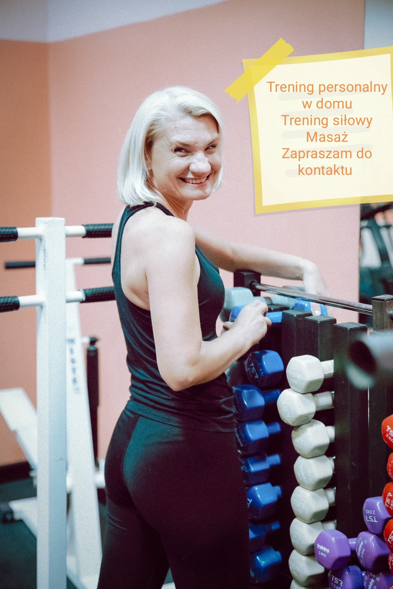 Uśmiechnięta kobieta w sportowym stroju stoi przy stojaku z hantlami w siłowni, na ścianie kartka z napisem 'Trening personalny w domu, Trening siłowy, Masaż, Zapraszam do kontaktu'.