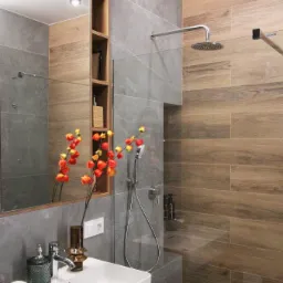 Nowoczesna łazienka z szarymi i drewnopodobnymi płytkami, białą umywalką nablatową, armaturą w kolorze chromu, bukietem miechunek i wnęką z półkami.