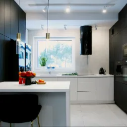 Nowoczesna kuchnia z czarnymi szafkami, białym blatem i wyspą, oświetleniem punktowym i wiszącymi lampami, na wyspie dekoracyjny talerz z owocami.