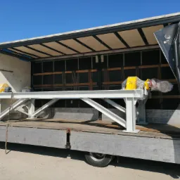 Biała, pomalowana konstrukcja stalowa z żółtymi elementami, owinięta folią, umieszczona na platformie ciężarówki z otwartą plandeką w słoneczny dzień.