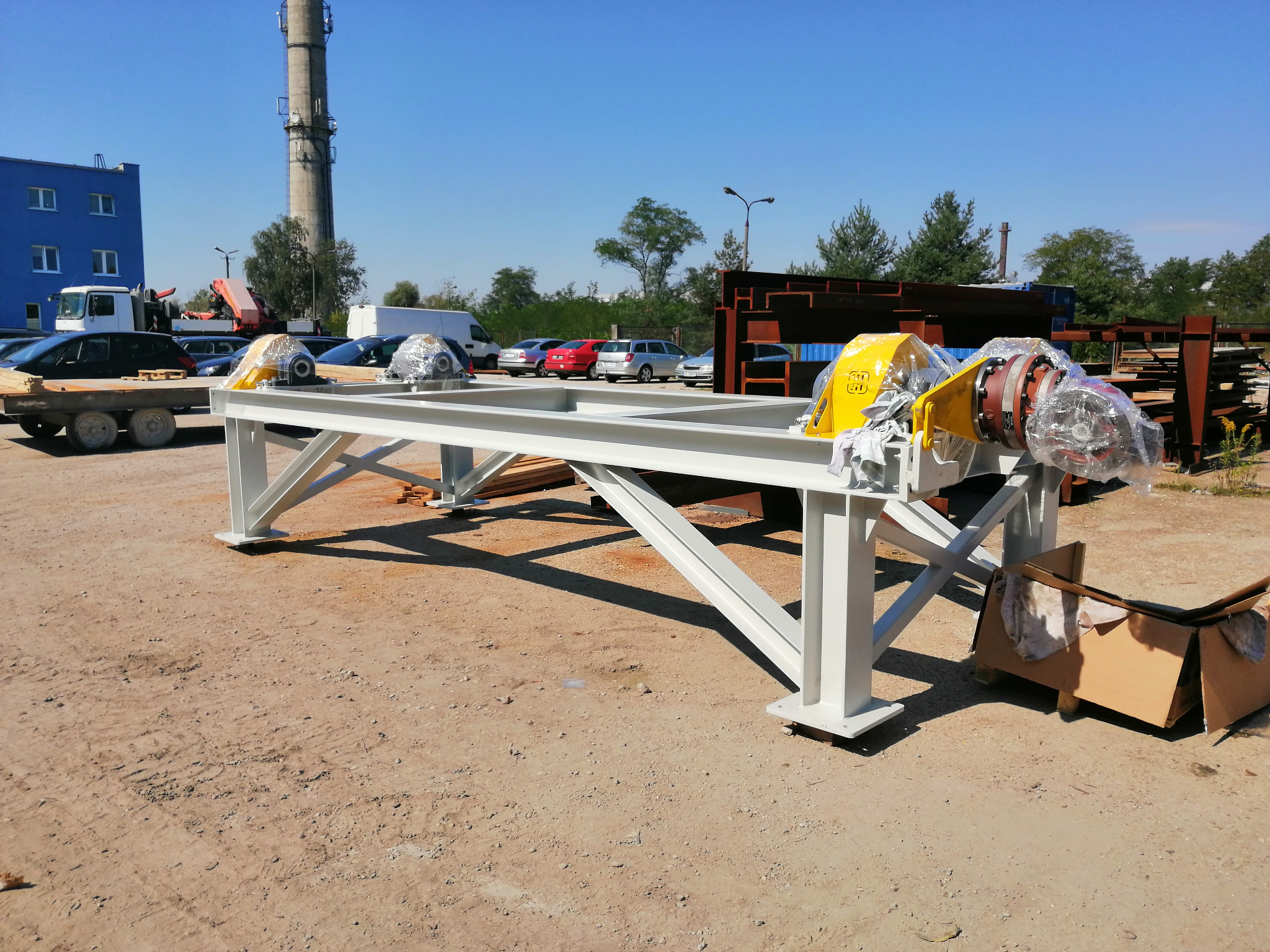Biała, pomalowana konstrukcja stalowa z dwoma żółtymi elementami przekładni i czerwonymi kołnierzami, zabezpieczonymi folią, na placu budowy z budynkiem i kominem w tle.