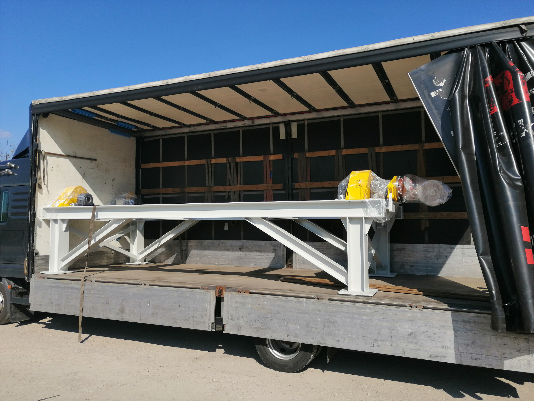 Biała, pomalowana konstrukcja stalowa z żółtymi elementami, owinięta folią, umieszczona na platformie ciężarówki z otwartą plandeką w słoneczny dzień.