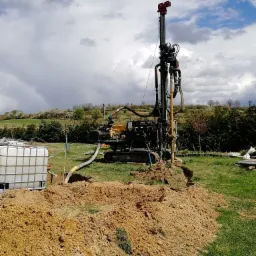 Wiertnica do studni głębinowej w trakcie pracy, widoczny zbiornik na wodę i wykopana ziemia, w tle zielony krajobraz z drzewami i pochmurne niebo.