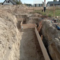 Szeroki kadr na wykop pod fundamenty z częściowo wykonanym szalunkiem z desek; w tle widoczny mężczyzna i zabudowa jednorodzinna.