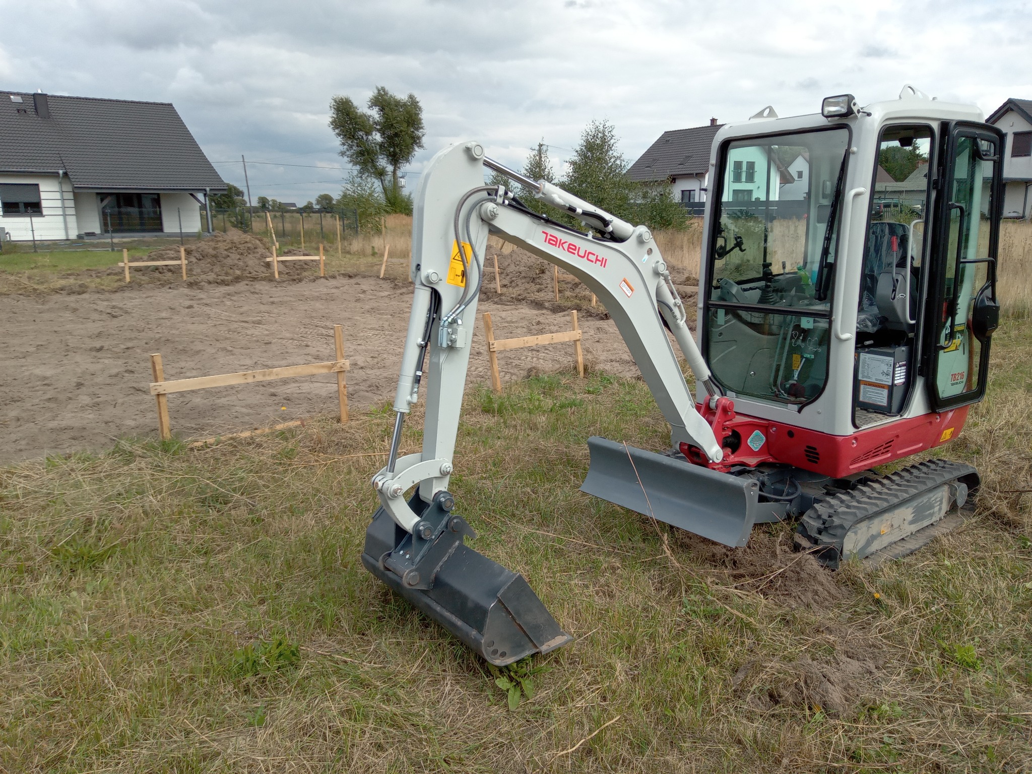 Kompaktowa koparka Takeuchi na trawiastym terenie, w tle wytyczony teren pod budowę fundamentów z drewnianymi kołkami i linkami, widoczne domy jednorodzinne, pochmurne niebo.